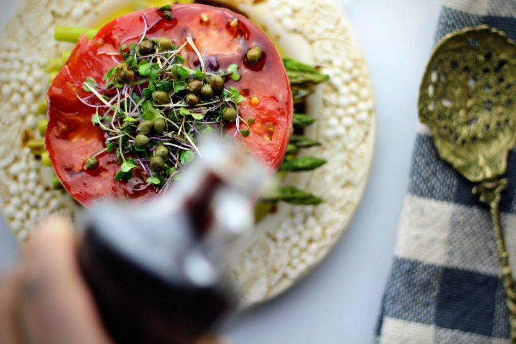 Pouring balsamic vinegar onto a stacked caprese garnished with microgreens and capers