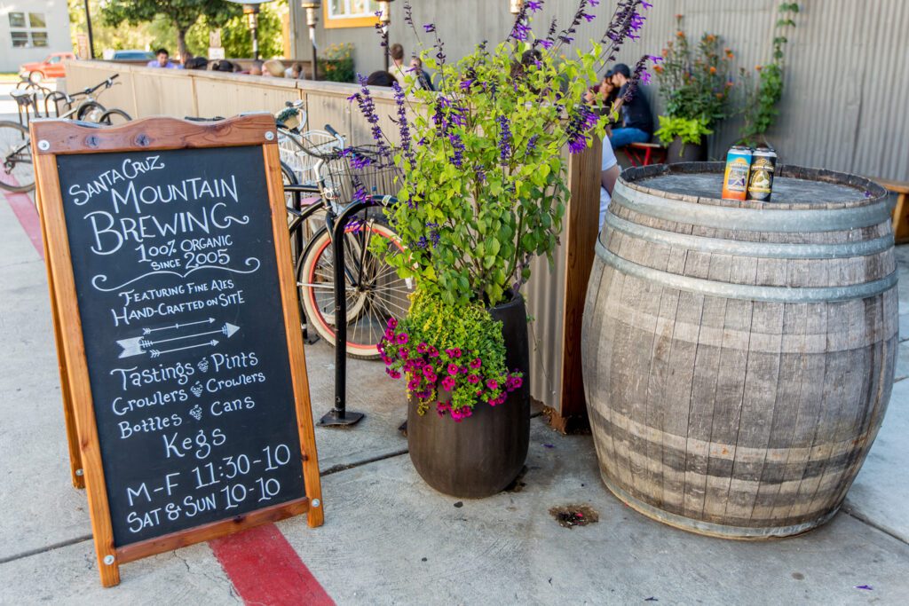 A sign, blooms, beer cans and barrel at the entrance to the beer garden at Santa Cruz Mountain Brewing