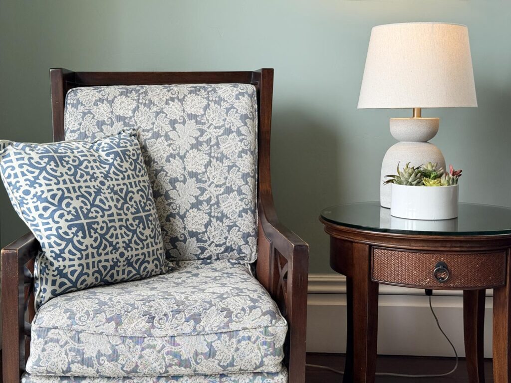 Close up photo of a upholstered chair with a pillow, end table, and table in the parlor at West Cliff Inn